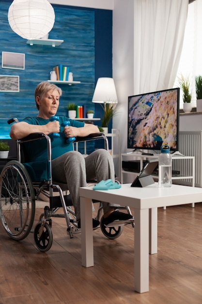 Donna anziana non valida in sedia a rotelle che allena i muscoli del corpo usando manubri ginnici