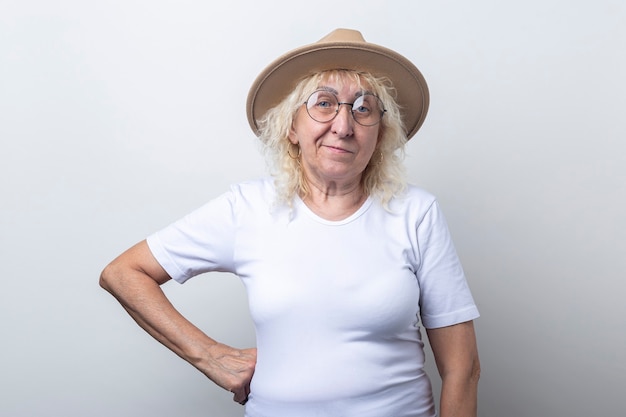 Donna anziana pensierosa in un cappello con gli occhiali su uno sfondo chiaro.