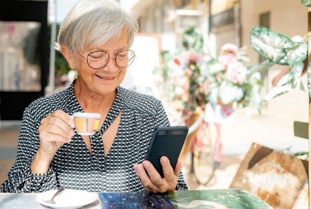 Donna anziana rilassata seduta al caffè all'aperto utilizzando il suo smartphone mentre si gusta una tazza di caffè espresso