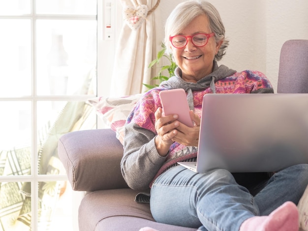 Donna anziana rilassata sul divano di casa utilizzando la tecnologia con computer portatile e smartphone. Luce intensa dalla finestra