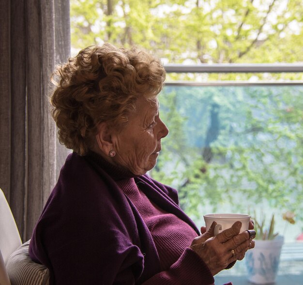 Donna anziana seduta vicino alla finestra con una tazza di caffè