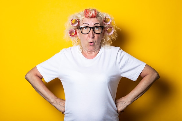 Donna anziana sorpresa con i bigodini in una maglietta bianca su una superficie gialla