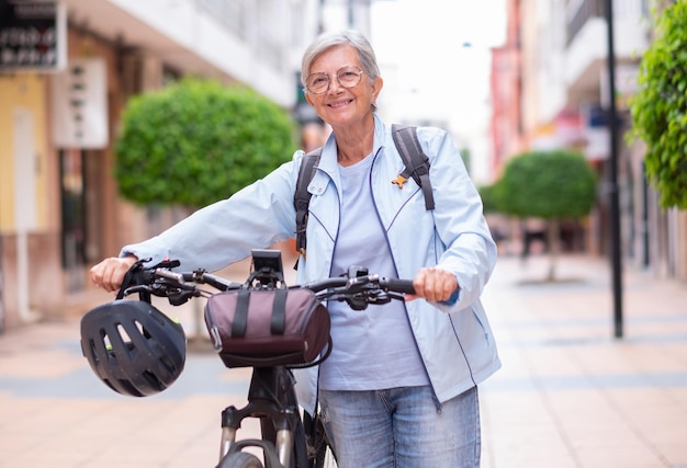 Donna anziana sorridente attraente del ciclista che spinge la sua bicicletta elettrica in una strada della città che esamina macchina fotografica Nonna attiva anziana che gode di uno stile di vita sano e del tempo libero in pensione