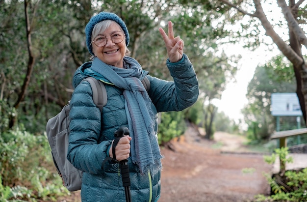 Donna anziana sorridente che cammina all'aperto in un sentiero di montagna godendosi la libertà e la natura Donna anziana allegra con zaino che fa un'escursione nel parco facendo un gesto positivo con le dita