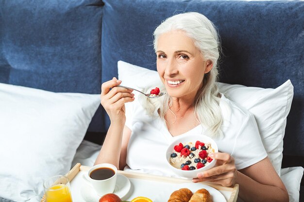 Donna anziana sorridente che mangia farina d'avena con frutti di bosco nel letto e guarda la telecamera
