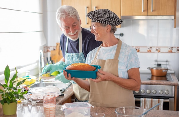 Donna anziana sorridente che mostra il suo plumcake fatto in casa appena sfornato mentre suo marito lava i piatti Concetto di famiglia e convivenza