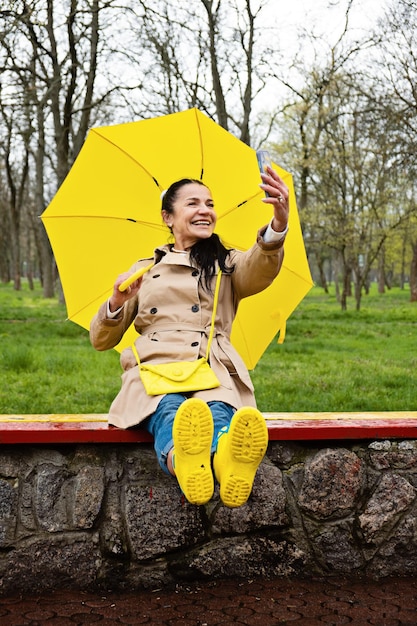 Donna anziana sorridente felice in impermeabile giallo con ombrello giallo che prende selfie sotto la pioggia primaverile