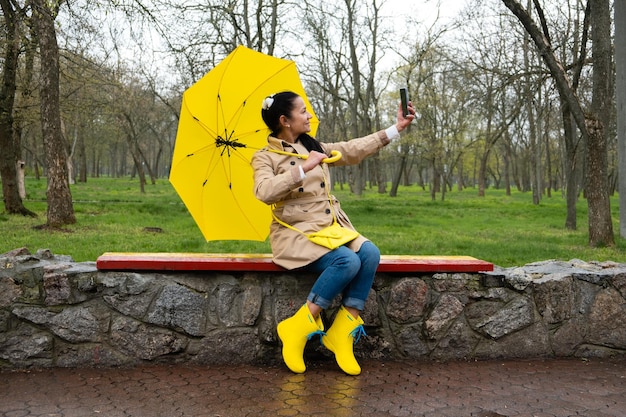 Donna anziana sorridente felice in impermeabile giallo con ombrello giallo che prende selfie sotto la pioggia primaverile