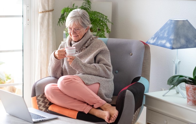 Donna anziana sorridente seduta a casa con una tazza di caffè che naviga sul laptop godendo di tecnologia e social media vecchia generazione e nuovo concetto tecnologico