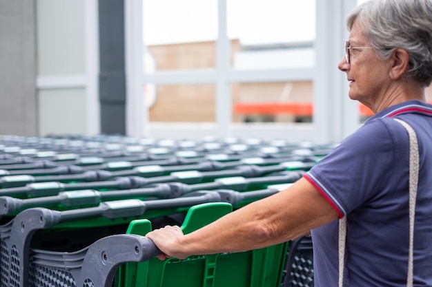 Donna anziana vicino a un gruppo di carrelli della spesa verdi vuoti fuori dal supermercato