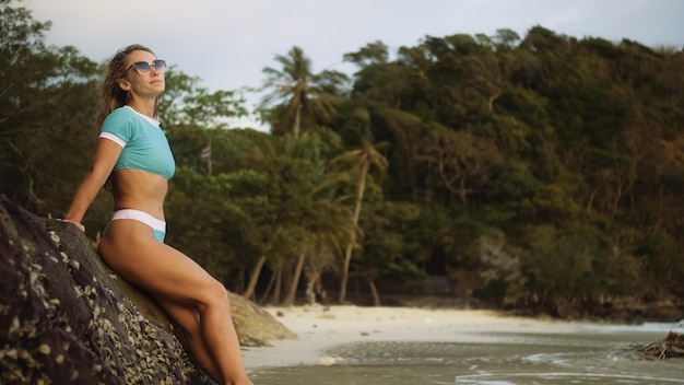 Donna appoggiata su una scogliera di roccia godendo e rilassarsi caldo tramonto dorato scuro Palma in spiaggia Donna in costume da bagno blu e occhiali da sole