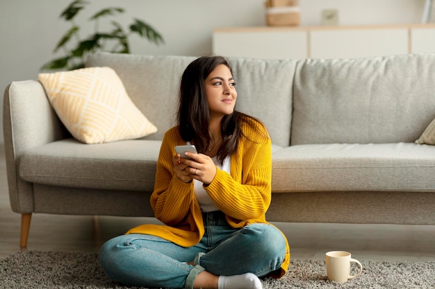 Donna araba sognante seduta sul pavimento con smartphone e tazza di caffè che lavora o studia da casa spazio libero