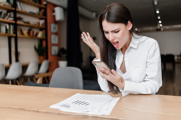 Donna arrabbiata in ufficio guardando lo schermo del telefono con documenti sulla scrivania