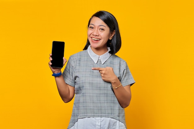 Donna asiatica allegra che mostra lo schermo vuoto dello smartphone e lo punta mentre guarda la telecamera su sfondo giallo