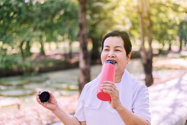 Donna asiatica anziana che beve acqua dopo la pausa che si esercita in natura, concetto di donna anziana in buona salute