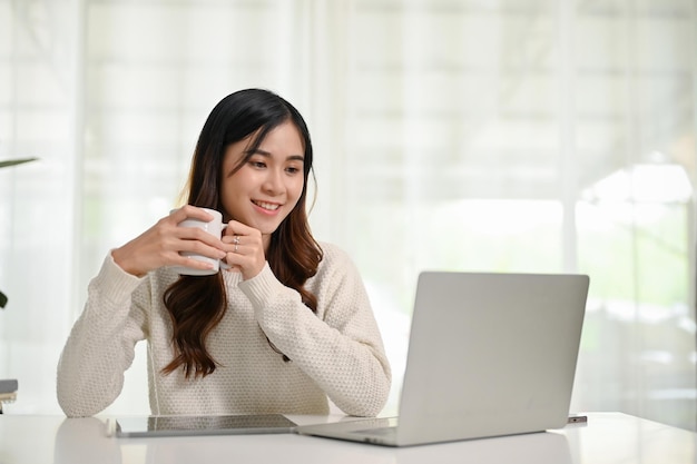 Donna asiatica attraente che tiene una tazza di caffè che esamina lo schermo del suo computer portatile