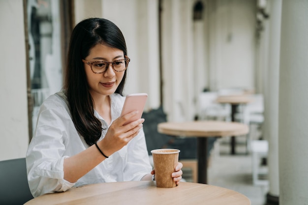 Donna asiatica attraente in un caffè di strada che legge un messaggio di testo dal suo smartphone seduto al tavolo all'aperto. giovane elegante signora dell'ufficio che si rilassa nell'ora del tè pomeridiano dal lavoro. ragazza felice con gli occhiali