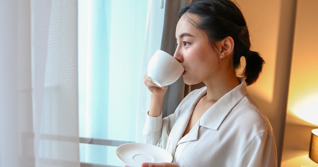 Donna asiatica che beve caffè e guardando fuori dalla finestra in casa.