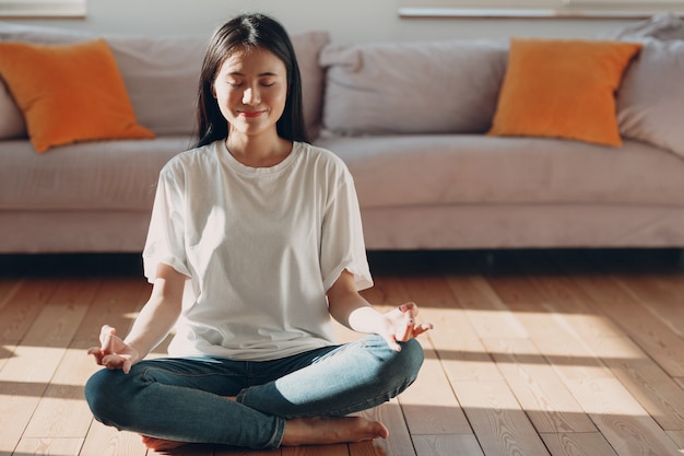 Donna asiatica che fa yoga e zen come la posa del loto meditativo in abbigliamento casual in un soggiorno al coperto a parte...