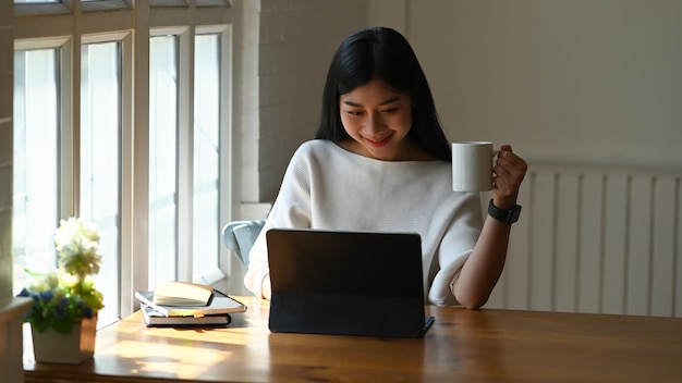 Donna asiatica che lavora il suo computer della compressa e che tiene una tazza da caffè in Ministero degli Interni.