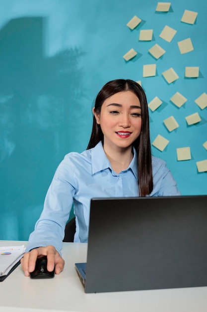 Donna asiatica che lavora su un computer portatile in un ufficio moderno che fa ricerche commerciali in una società di avvio. Donna professionale seduta alla scrivania utilizzando il mouse wireless concentrandosi sul compito.