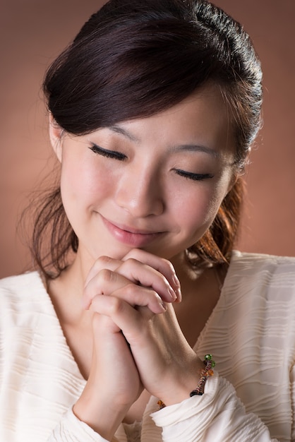 Donna asiatica che prega, ritratto del primo piano su fondo marrone dello studio.
