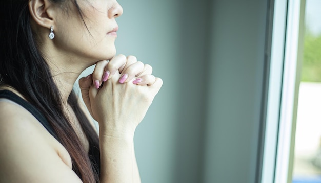 Donna asiatica che prega vicino alla finestra nella stanza Mani giunte nel concetto di preghiera per la fede, la spiritualità e la religione