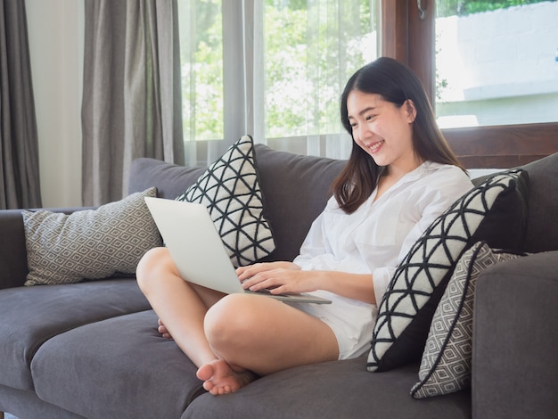 Donna asiatica che si siede e che utilizza computer sul sofà a casa