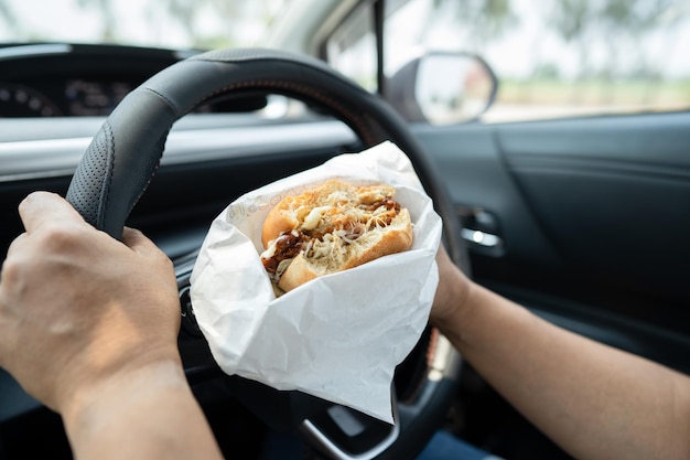 Donna asiatica che tiene un hamburger e delle patatine fritte da mangiare in macchina pericolosa e rischia un incidente