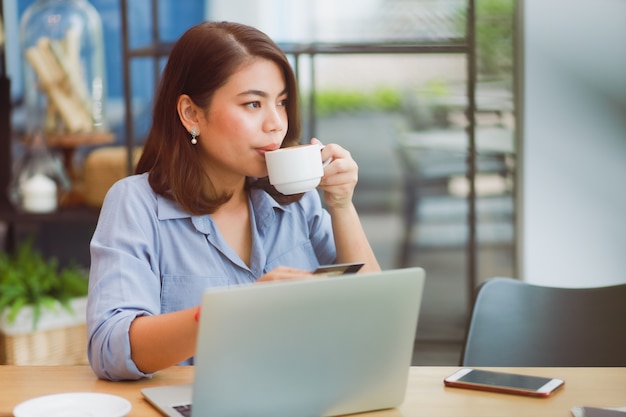 Donna asiatica che utilizza telefono cellulare con la carta di credito e il computer portatile per il pagamento online di compera nel caffè della caffetteria con gli amici