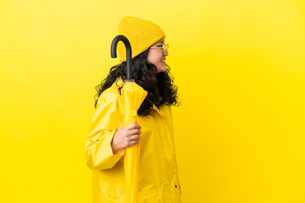 Donna asiatica con cappotto antipioggia e ombrello isolato su sfondo giallo che ride in posizione laterale