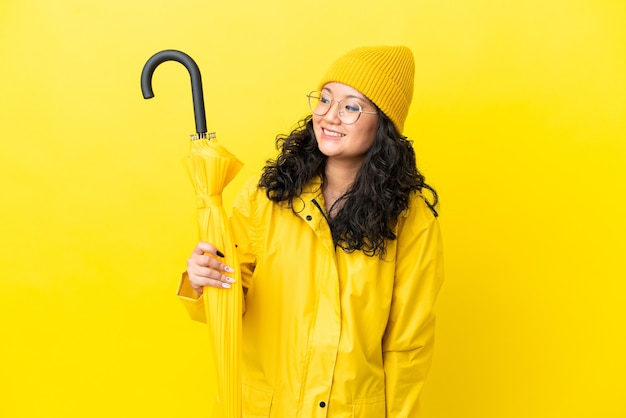 Donna asiatica con cappotto antipioggia e ombrello isolato su sfondo giallo guardando di lato e sorridente