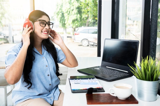 Donna asiatica con gli occhiali che ascolta la musica con le cuffie con un computer portatile e un taccuino con un telefono cellulare e una tazza di caffè sul tavolo