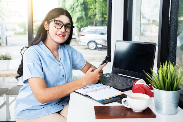 Donna asiatica con occhiali utilizzando un telefono cellulare con un computer portatile e un taccuino con una tazza di caffè sul tavolo