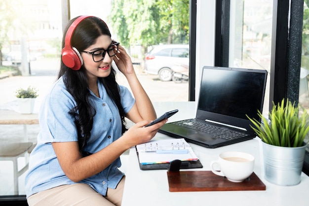 Donna asiatica con occhiali utilizzando un telefono cellulare e cuffie con un computer portatile e un taccuino con una tazza di caffè sul tavolo