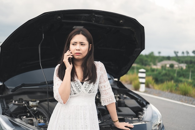 Donna asiatica con un&#39;automobile rotta che per mezzo del telefono cellulare per chiamare assistenza sul bordo della strada.