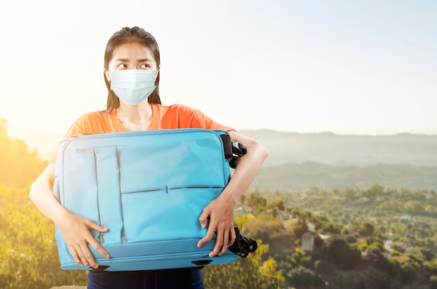 Donna asiatica con una maschera facciale che trasporta con una valigia sul campo. Viaggiare nella nuova normalità