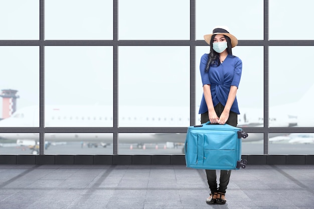 Donna asiatica con una maschera facciale con una valigia al terminal dell'aeroporto. In viaggio nella nuova normalità