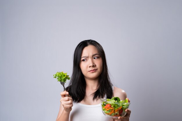 Donna asiatica confusa con il mangiare insalata isolata sopra il muro bianco. Stile di vita sano con il concetto di cibo pulito.