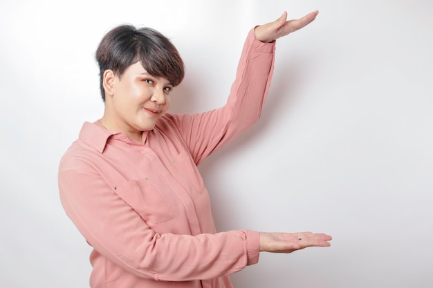 Donna asiatica dai capelli corti eccitata che indossa una camicia rosa che punta allo spazio della copia accanto a lei isolato da uno sfondo bianco