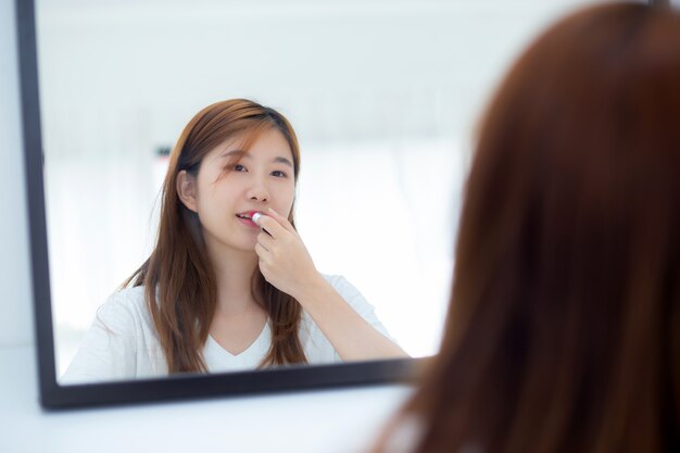 Donna asiatica del ritratto di bellezza allo specchio che tiene e che guarda un rossetto di trucco