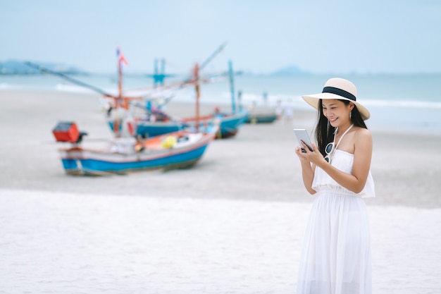 Donna asiatica del viaggiatore che per mezzo del telefono cellulare sulla spiaggia