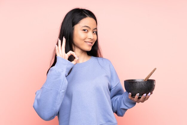 Donna asiatica dell'adolescente isolata sul beige che mostra segno giusto con le dita mentre tiene una ciotola di tagliatelle con le bacchette