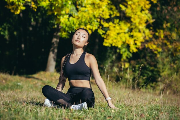 Donna asiatica della giovane bella ragazza sportiva che medita nel parco, posa del loto seduto praticando la stuoia di yoga, zen. si rilassa all'aperto nella natura al mattino Concetto di stile di vita sano, relax, calma, meditazione