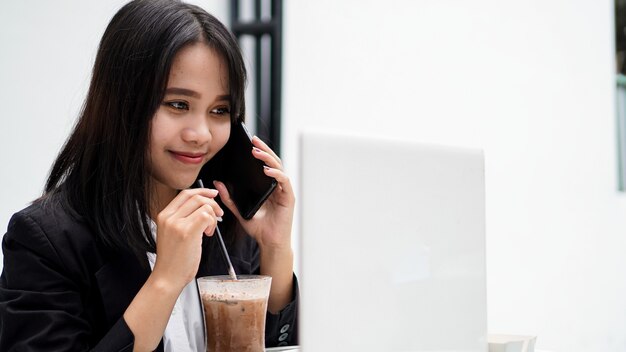 Donna asiatica di affari che dringking caffè e parlando sullo smartphone al caffè