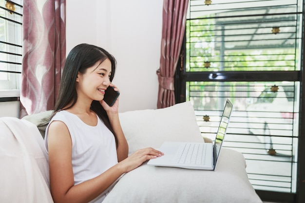 Donna asiatica di affari che lavora con il telefono cellulare e il computer portatile sul divano di casa