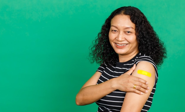 Donna asiatica di mezza età che tiene il braccio con una benda che mostra che è stata vaccinata per il virus Covid 19 su sfondo verde. Concetto per la vaccinazione contro il Covid 19.