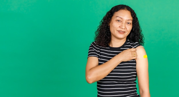 Donna asiatica di mezza età che tiene il braccio con una benda che mostra che è stata vaccinata per il virus Covid 19 su sfondo verde. Concetto per la vaccinazione contro il Covid 19.