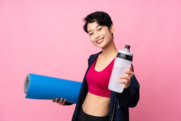 Donna asiatica di sport giovane sopra la parete rosa isolata con la bottiglia di acqua di sport e con una stuoia