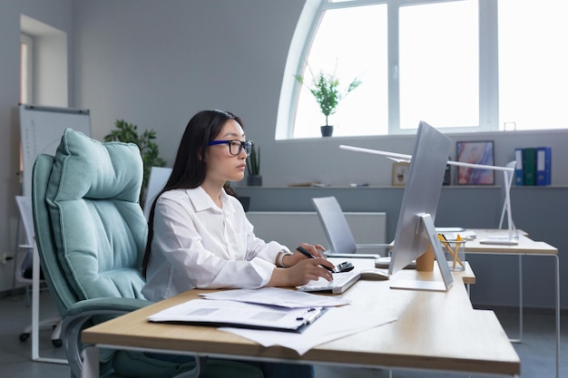 Donna asiatica di successo di affari che lavora con il computer nell'ufficio moderno premuroso e concentrato